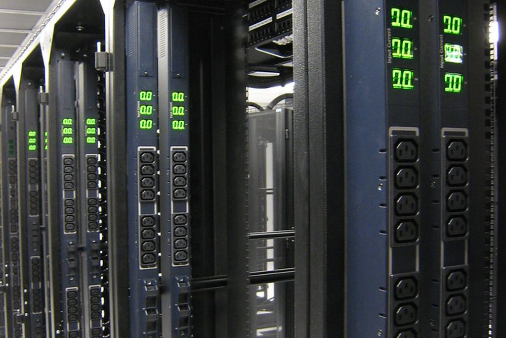 Server racks in a dark data hall, power meters glowing green. Photograph by bugeaters on Flickr.